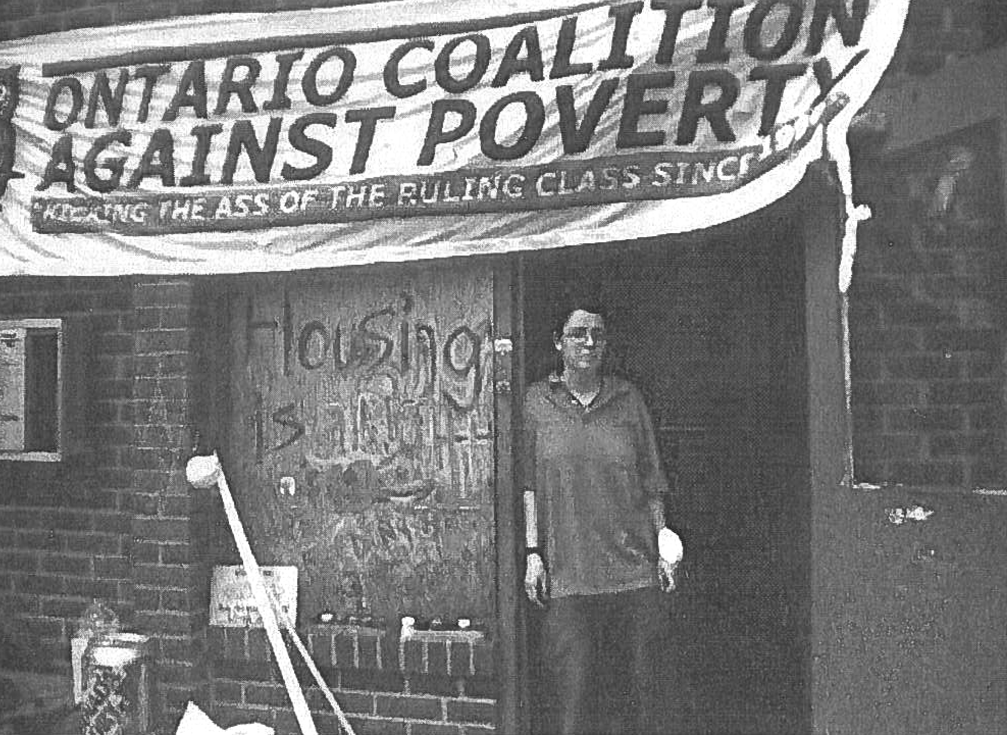black and white photograph with a person standing in a doorywau with a banner over their head which reads Ontario Coalition Against Poverty Kicking the ass of the ruling class since 19... 
